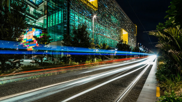 Image at night of tram tracks with a building in the background lit up with green lights. The are blue and red light beams above the tracks also giving it a sense of movement
